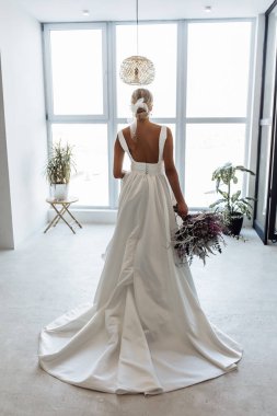 Beyaz elbiseli, güzel bir düğün buketi ile modern iç mekanda poz veren çekici genç gelinin tam boy portresi.                   