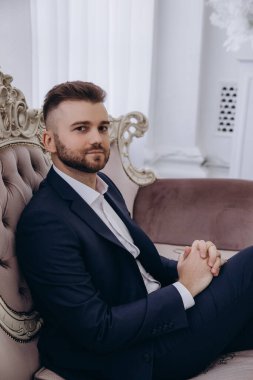 Şık takım elbiseli yakışıklı genç adam koltukta oturuyor ve kameraya bakıyor.