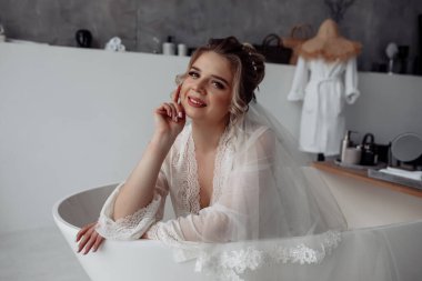 Güzel, mutlu, beyaz cüppeli, duvak takmış, banyoda poz veren genç bir gelin.                 