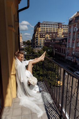 Beyaz cüppeli, duvak takmış, şehirde balkonda poz veren çekici bir gelin.            