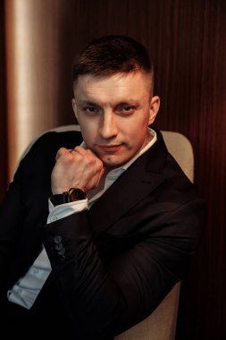 Siyah takım elbiseli genç ve çekici bir adam koltukta oturmuş kameraya bakıyor.