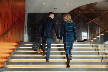 Modern otel koridorunda el ele tutuşup merdivenleri çıkan genç bir çift.  