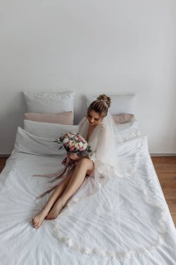 Beyaz cüppeli, duvak takmış, sabah yatak odasında poz veren güzel gelinin portresi.         