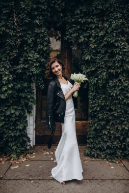 Gelinlik ve deri ceketli gelin. Parkta kameraya gülümseyen bir buket çiçekle.