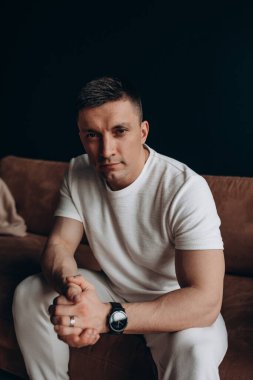 Kanepede beyaz tişört giyen ciddi yüzlü yakışıklı bir genç.