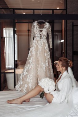 Mutlu genç gelin elinde buket ve gelinliğin yanında yatağında oturuyor.