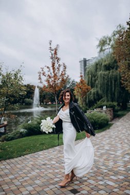Beyaz elbiseli, deri ceketli, elinde düğün buketi olan ve dışarıda poz veren güzel bir gelin.             