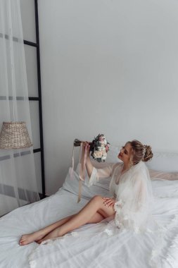 Mutlu genç gelin bir buket çiçekle yatakta oturuyor.