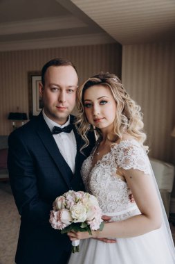 Gelin ve damat birlikte poz verip odada kameraya bakıyorlar.