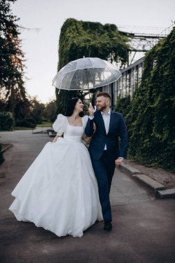 Gelin ve damat parkta şemsiyeyle yağmurda yürüyorlar