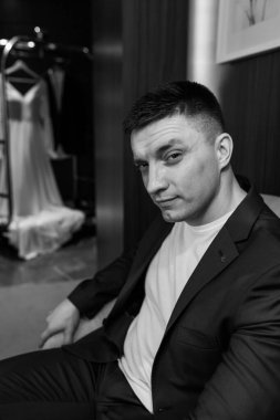 Siyah beyaz takım elbiseli yakışıklı bir genç adam oturuyor ve kameraya bakıyor.