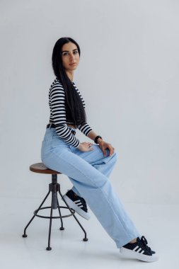 Genç esmer kadın sandalyede oturuyor ve gri arka plandaki kameraya bakıyor.