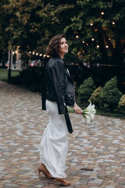 Beyaz elbiseli, siyah deri ceketli ve dışarıda kameraya poz veren beyaz bir gelin. 