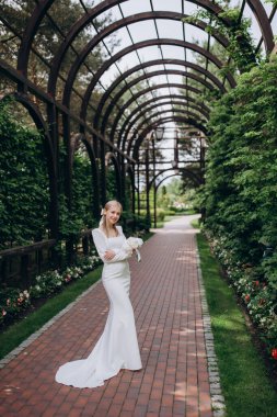 Yeşillik ve çalılıkların arkasında yürüyen gelinlik içinde mutlu genç bir gelin.