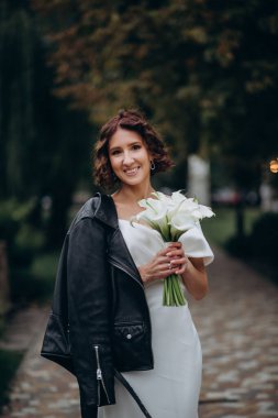Beyaz elbiseli, siyah deri ceketli, açık havada poz veren esmer kadın. 