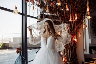 Beyaz gelinlik ve duvak giymiş güzel genç gelinin portresi rahat iç mekânda poz veriyor.