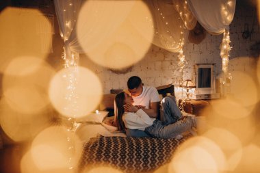 Kafkasyalı mutlu çift, hoş bir otel odasında çelenk ışıkları ve bokeh efektiyle vakit geçiriyor. 