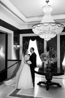 Gelin ve damat zarif iç mekanda birlikte poz veriyorlar, siyah ve beyaz.