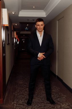 Takım elbiseli yakışıklı adam otel koridorunda kameraya gülümsüyor.