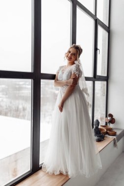 Gelin beyaz elbiseli ve peçeli pencerenin yanında duruyor ve kameraya gülümsüyor.