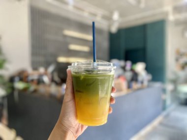 Bir kafede plastik bir bardak Matcha Yuzu tutan kadın eli. Matcha Yuzu altta yuzu suyu, üstte de kibrit. Arka planda bulanık bir görüntü..
