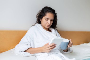  Young woman drinking coffee in bed in bathrobe reading an ebook