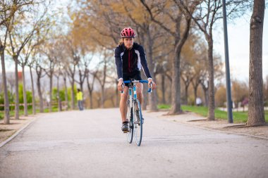 Spor giyim ve kask takan yakışıklı bir bisikletçi parktaki yolda bisiklet sürüyor.
