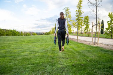 Çıplak ayaklı, spor kıyafetli, patenli, parkta yürüyen genç bir erkeğin arka görüntüsü.