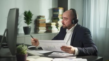 Kulaklıklı bir işadamı online bir toplantıda grafikler gösteriyor. Adam evden çalışıyor ve ortaklarıyla video bağlantısı üzerinden konuşuyor, evrakları gösteriyor ve iş konularını tartışıyor. Yüksek kalite 4k görüntü