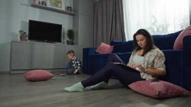 Bir kadın bilgisayarda çalışıyor ve oğlu oturma odasında oyuncaklarla oynuyor. Evden internet üzerinden çalışacağım. Aile bütün günü odada birlikte geçirir. Yüksek kalite 4k görüntü