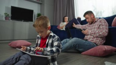 Çocuk tabletle oynarken ebeveynler evde bilgisayar ve telefonla çalışırlar. Oturma odasındaki işle ilgili online toplantılar ve telefon aramaları. Ebeveynler meşgulken çocuk oynuyor.. 