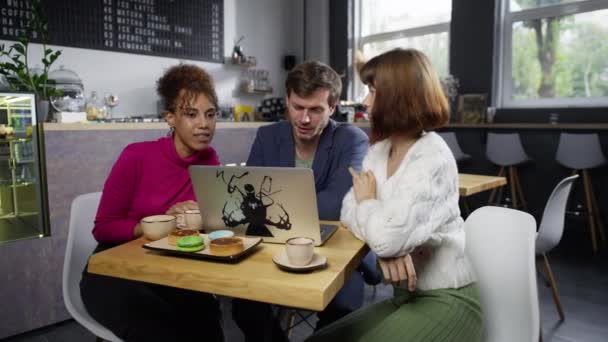 カフェのコンピュータでの学生のオンライン会議 男と二人の多国籍の女の子がコーヒーを飲んでいて ウェブカメラでチャットしている 高品質4K映像 — ストック動画