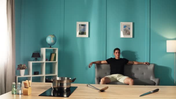一个长相像欧洲人的男人正在沙发上休息 而一顿饭则在炉子上做饭 一个人在家做饭 这个人在休息 高质量的4K镜头 — 图库视频影像