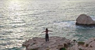 Aerial view of a happy woman at sunset on the rocks near the sea. The girl has melted her hands and feels freedom on the ocean shore. High quality 4k footage