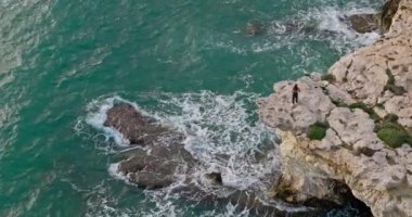 Aerial view of a young woman standing on the rocks on the ocean shore. A beautiful girl alone looks at the sea and waves in a beautiful landscape. Ludany and the amazing nature of the ocean, mountains