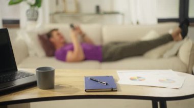 Man resting on the couch using the phone with social networks after a hard days work. Home office with a computer and documents on the table. Handsome Caucasian guy at home with a smartphone. 