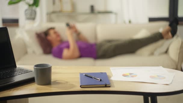 男は仕事の後 ソーシャルネットワークと電話を使用してソファの上で休んでいます テーブルの上にコンピュータとドキュメントを持つホームオフィス ハンサムな白人男性とともに自宅でスマートフォン — ストック動画