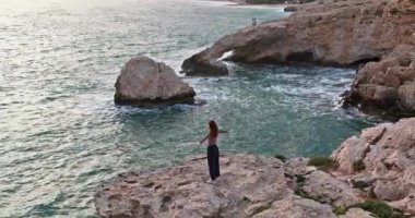 Aerial view of a happy woman on the edge of the rocks on the seashore with her arms crossed. The girl feels freedom and unity with nature. Beautiful ocean landscape of waves and stones.