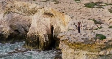 Aerial view of a woman standing on the edge of a rocky cliff on the seashore. Little man on the ocean shore with big waves. A girl on the edge of nature. High quality 4k footage