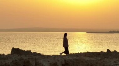 Deniz kıyısı boyunca gün ışığında yürüyen bir kadın siluetinin havadan görünüşü. Güzel, romantik bir kız, güneşin altında okyanus kıyısında rahatlık ve mutluluk hisseder. Yüksek kalite 4k 