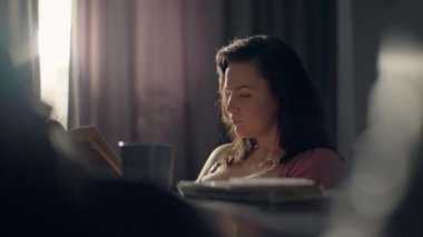 Avrupalı kız kiga okuyor ve evde sabah kahvesi içiyor. Sabah atmosferinin sade sıcaklığı kavramı. Kitapların yardımıyla bir kadının eğitimi. Yüksek kalite 4k görüntü