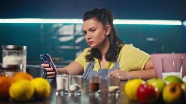 Bir kadın telefondaki yemek tarifi ile ürünleri karşılaştırır. Mutfaktaki kız akşam yemeğini hazırlamaya hazır. Yüksek kalite 4k görüntü
