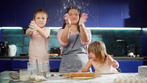 息子と娘を持つ母親がキッチンで生地を準備しています 家族は台所で楽しんでいて 小麦粉を広げています 一緒に幸せな子供たちは料理に役立ちます 高品質4K映像 — ストック動画