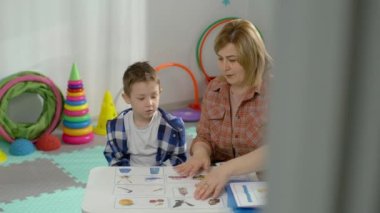 Öğretmen çocuğa resimlerle görevleri doğru şekilde nasıl yapacağını gösterir. Okulda oyun öğrenme ve geliştirme. Oğlan sınıftaki masada ders çalışıyor. Yüksek kalite 4k görüntü