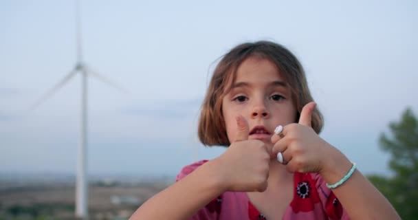 風車を背景に指を上げて姿を現す赤ん坊の女の子のクローズアップ 風からの環境保護とクリーン生態電力の概念 — ストック動画
