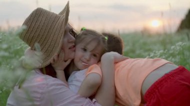 Çocuklar gün batımında çimenlerde otururken annelerine sıkıca sarılırlar. Aile mutluluğu kavramı ve oğul ve kız için anne sevgisi gülümseme ve yaşam tarzı. Yüksek kalite 4k görüntü