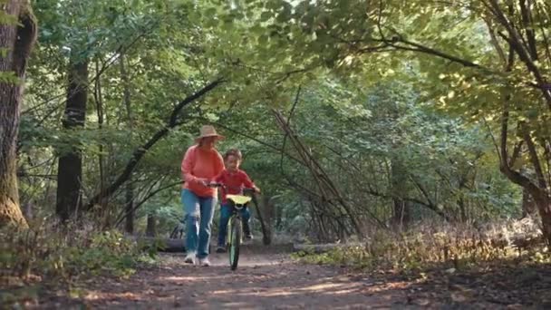 母親は息子に森で自転車に乗るように教える 女性は歩いている子供の自転車を支えています ペアレンタルケア 高品質の4K映像 — ストック動画
