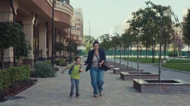 Sırt çantalı bir okul çocuğu şehir caddesi boyunca annesiyle birlikte okula koşar. Çocuk ders için aceleyle sınıfa gider. Yüksek kalite 4k görüntü