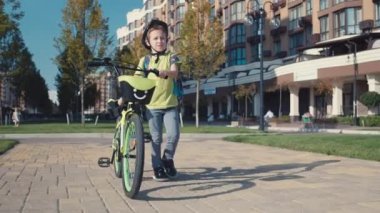 Omuzlarında sırt çantası ve bisikleti olan bir çocuk ders için okula gider. Aktif mutlu bir çocuk okula bisikletle koşuyor. Yüksek kalite 4k görüntü
