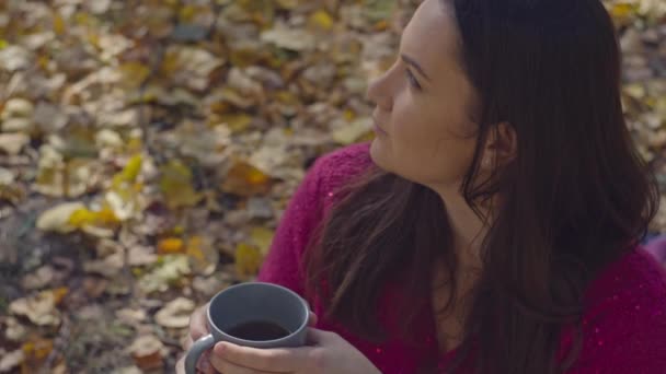 秋の公園で紅茶を飲むロマンチックな女性のクローズアップ 夢見る女の子は 黄金の季節に葉の上に座っている暖かいコーヒーを飲みます 高品質の4K映像 — ストック動画